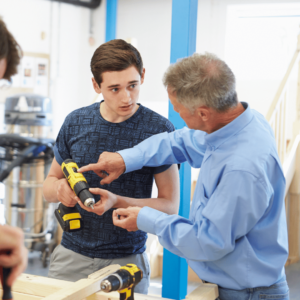 A carpentry teacher showing a student how to use a power drill